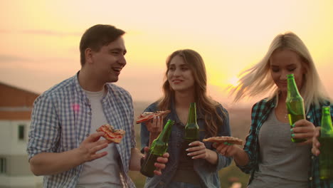 Loving-couple-drinks-beer-and-eats-hot-pizza-on-the-roof.-Girl-gently-puts-her-head-on-her-boyfriend's-shoulder.-They-are-smile-and-enjoy-the-time-at-the-sunset.